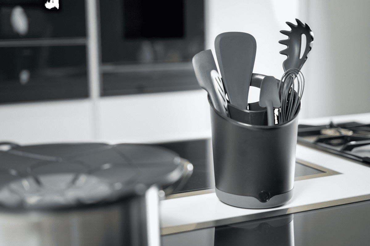 Ein eleganter schwarzer Utensilienbehälter der Firma Kochblume GmbH, bestückt mit unverzichtbaren Küchenutensilien, steht auf einer makellos weißen Arbeitsplatte neben einem modernen Kochfeld und einem Topf und verleiht Ihrem Küchenbereich sowohl Ordnung als auch zeitgemäßen Stil.