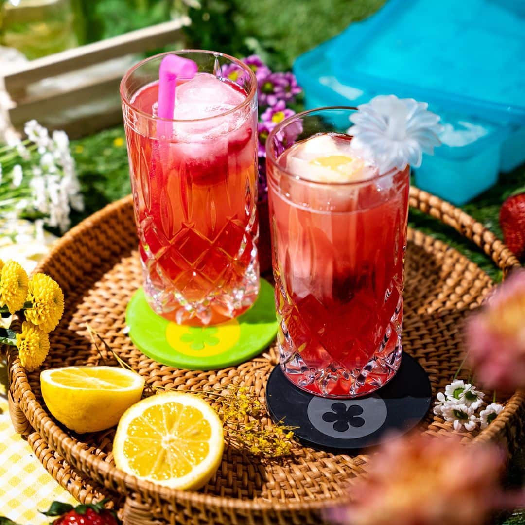 Zwei Gläser mit gekühlten Rotweingetränken, mit Strohhalmen und Obst, auf einem Weidenkorb mit Blumen und Zitronenhälften.