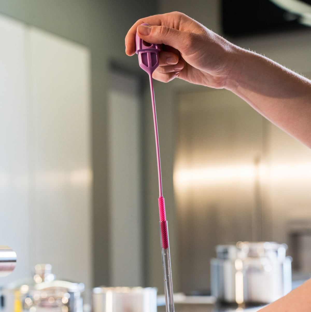 In einer modernen Küche hält eine Hand eine lange, violette Becherbürste der Firma Kochblume GmbH über einen Glasbehälter.