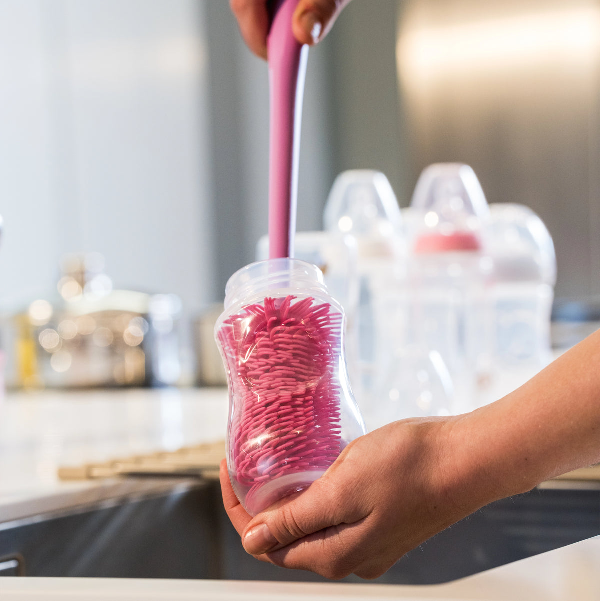 Eine Person benutzt eine rosa Becherbürste der Firma Kochblume GmbH, um das Innere eines Babyfläschchens am Waschbecken zu reinigen, während im Hintergrund weitere Fläschchen sorgfältig gereinigt werden.