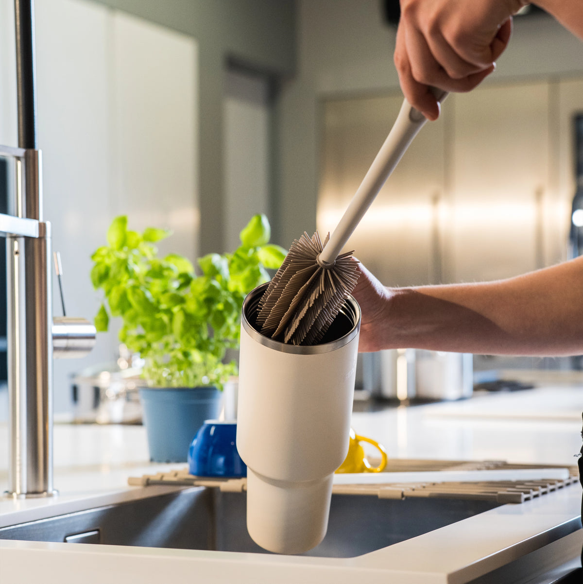 Eine Person reinigt mit einer Becherbürste der Firma Kochblume GmbH einen wiederverwendbaren Kaffeefilter in einem Spülbecken, neben dem eine Pflanze steht.