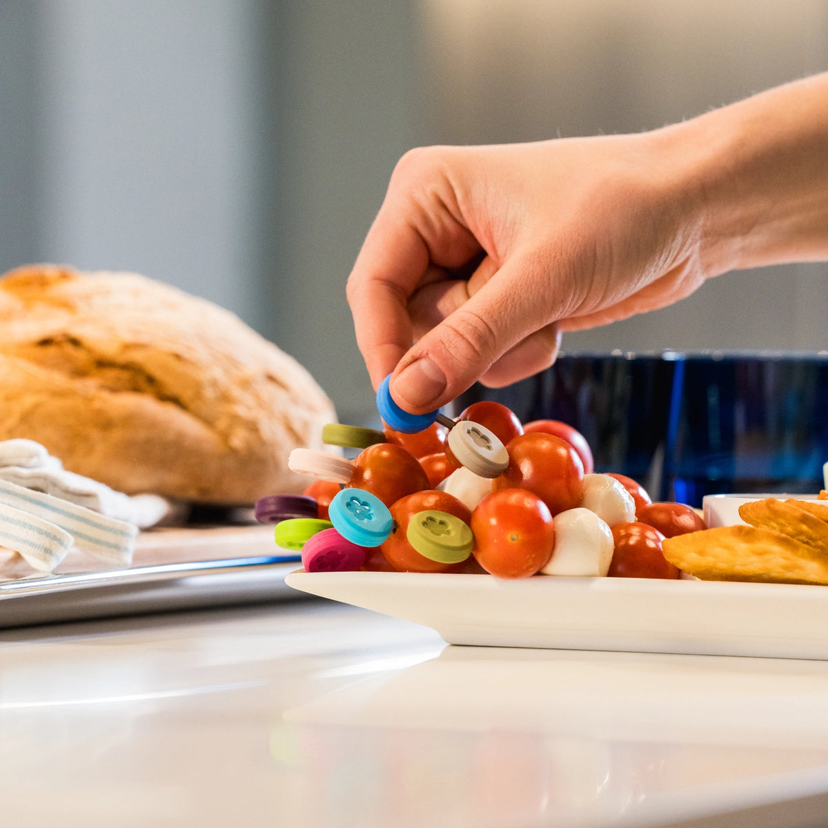 Sur le plan de travail de la cuisine, une main prend une « Snackgabel » colorée en forme de bouton, issue du lot de 10 « Snackgabel » de la société Kochblume GmbH, posée sur une assiette contenant des tomates cerises et des amuse-bouches.