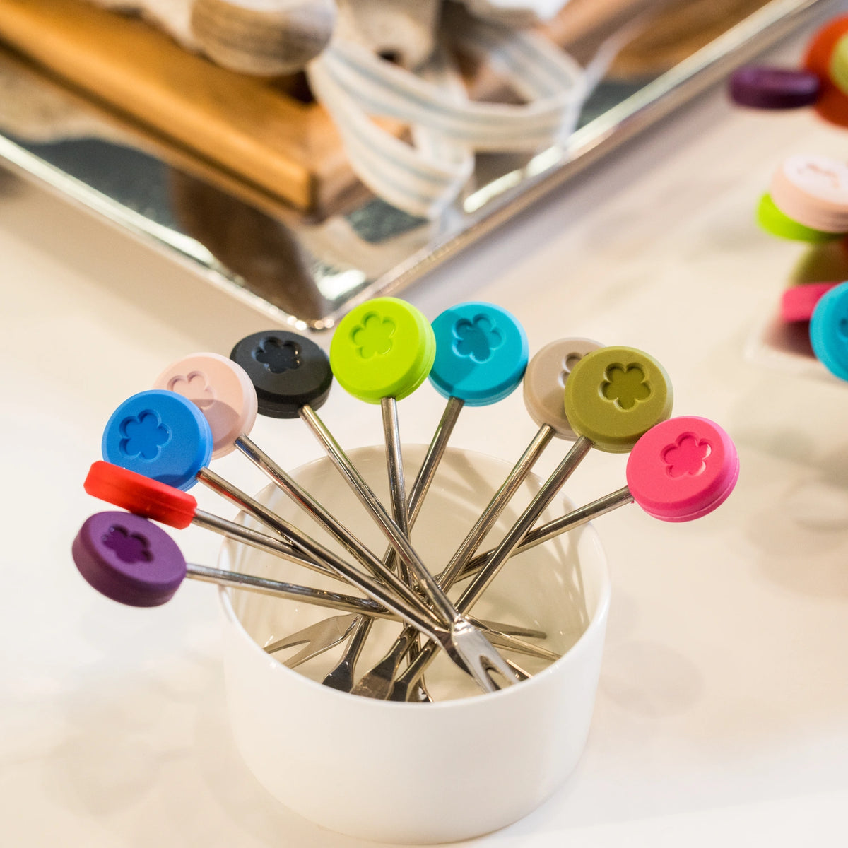 Kochblume GmbH, lot de 10 « Snackgabel » : des pics colorés en forme de fourchette fixés à des pinces métalliques, présentés aux côtés d'aimants en forme de boutons dans un gobelet blanc posé sur un plan de travail de cuisine.
