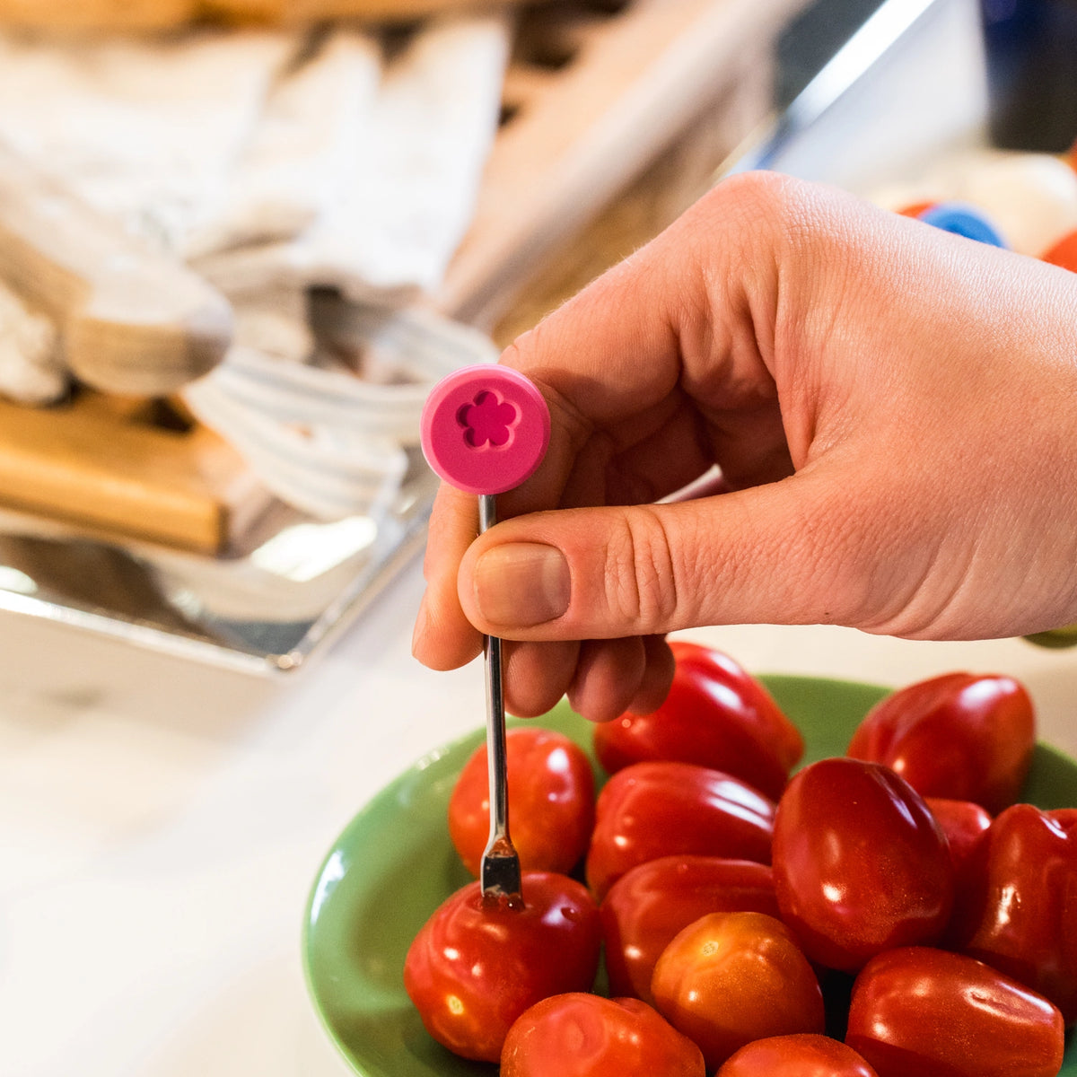 Une main utilisant un petit outil métallique à manche rose, issu du lot de 10 « Snackgabel » de la société Kochblume GmbH, pour percer des trous dans des tomates cerises placées dans un bol vert.