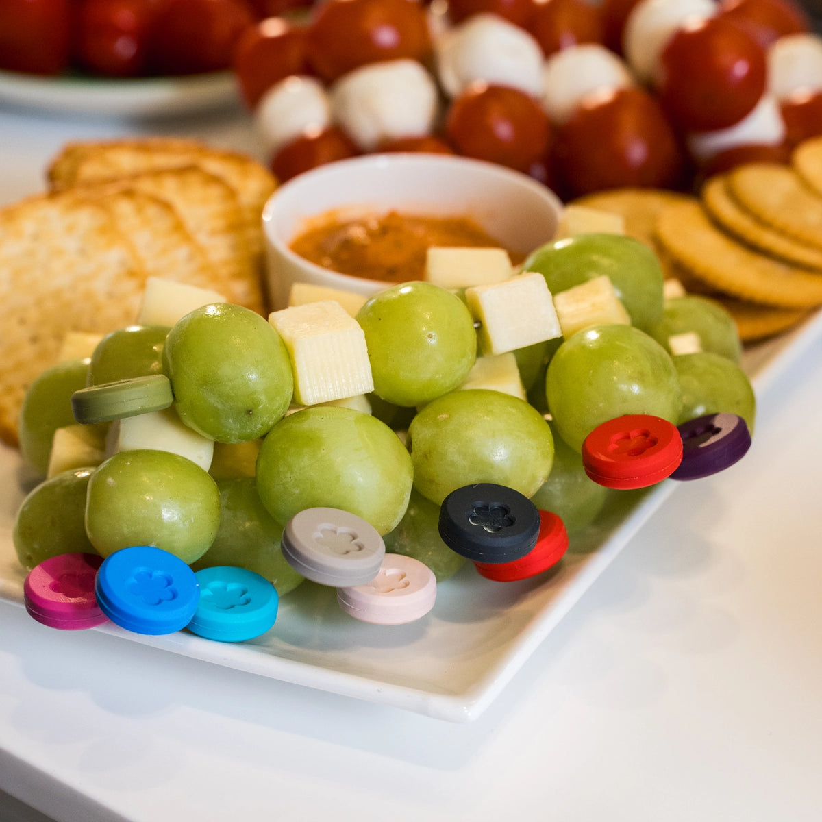 Des brochettes de raisins verts et de fromage, présentées sur une assiette avec les manches colorés du lot de 10 « Snackgabel » de Kochblume GmbH, avec des crackers en arrière-plan.