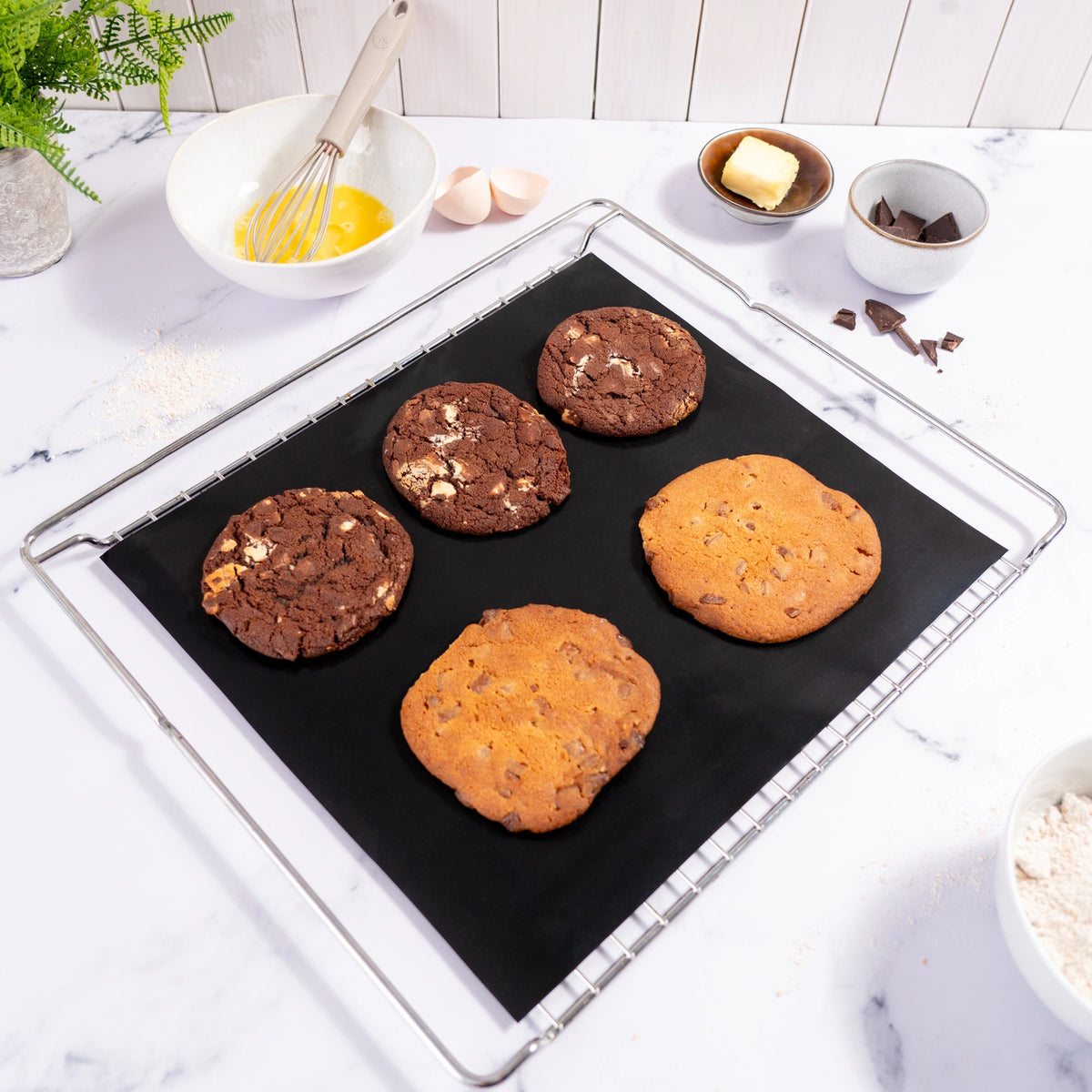 Four cookies—two chocolate and two chocolate chip—rest on a baking tray lined with the Dauerbackfolie 2tlg. by Kochblume GmbH, with baking ingredients arranged nearby.