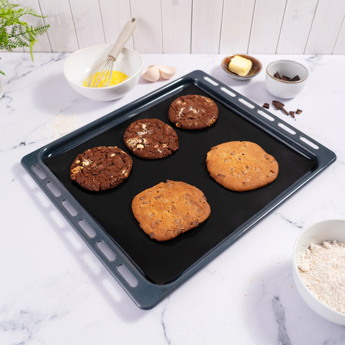 A Dauerbackfolie 2tlg. baking mat set from Kochblume GmbH is shown on a marble countertop, holding four freshly baked cookies—two chocolate and two plain—amidst an array of baking ingredients. This inviting scene perfectly showcases the durability and versatility of Kochblume GmbH’s Dauerbackfolie, making it an ideal choice for home bakers who want reliable results with every batch.