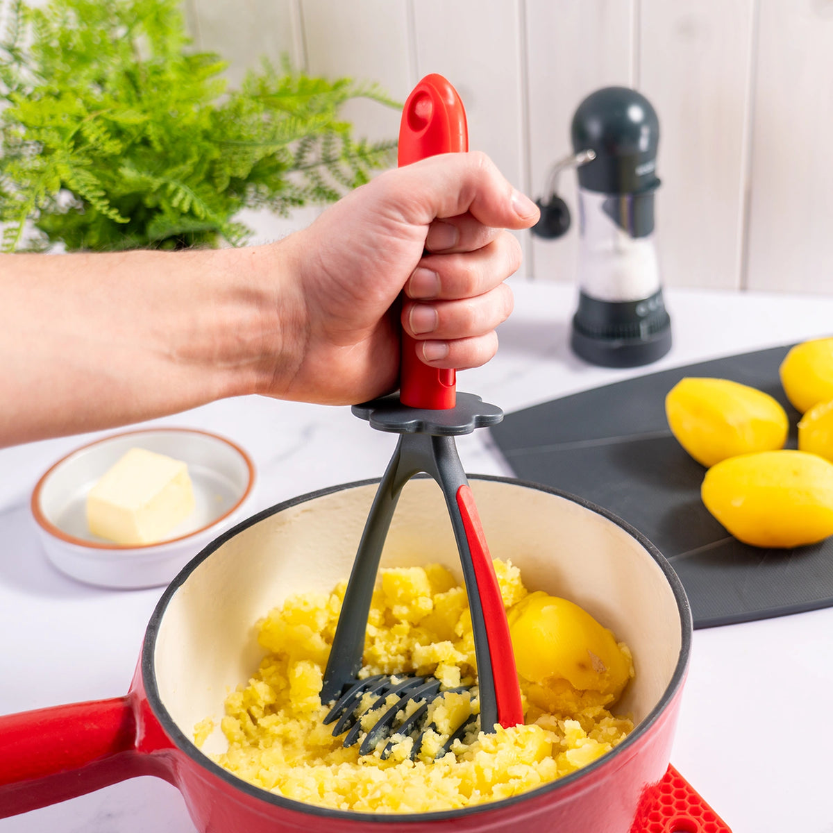 Eine Hand zerdrückt mit dem Kartoffelstampfer der Kochblume GmbH, einem roten Kartoffelstampfer, gekochte Kartoffeln in einem Topf auf einer Küchenarbeitsplatte. Es wird kein eigentlicher Produktbeschreibungstext angezeigt – es wird lediglich CSS-/Stilcode angezeigt.
