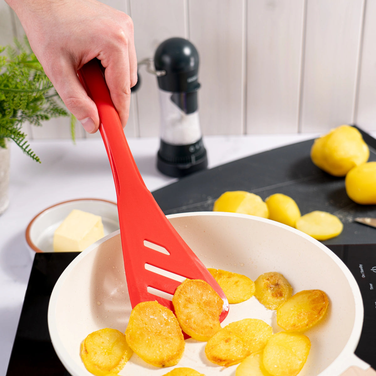 Eine Hand verwendet den Design-Schlitzwender der Kochblume GmbH – einen roten Schlitzwender –, um Kartoffelscheiben in einer weißen Pfanne auf einem Herd zu braten.