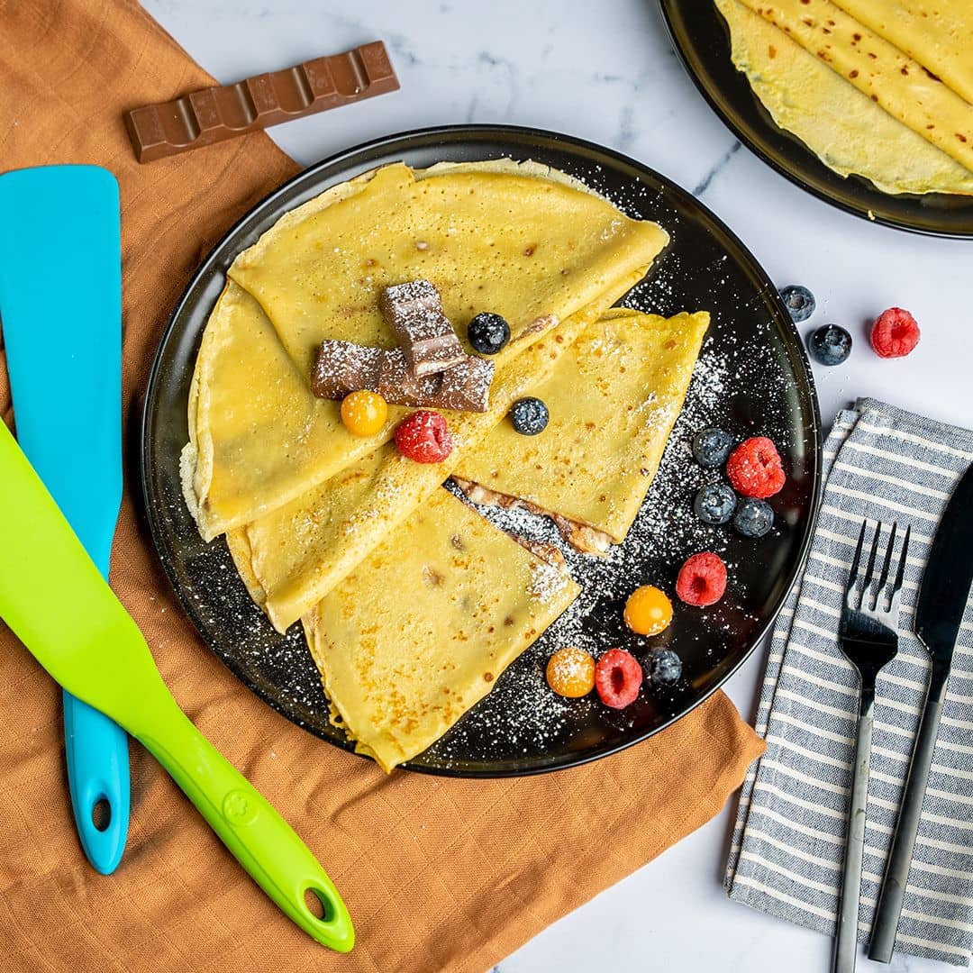 Ein Teller mit Crêpes mit Beeren, Schokoladenstückchen und Puderzucker, daneben Besteck und bunte Pfannenwender.