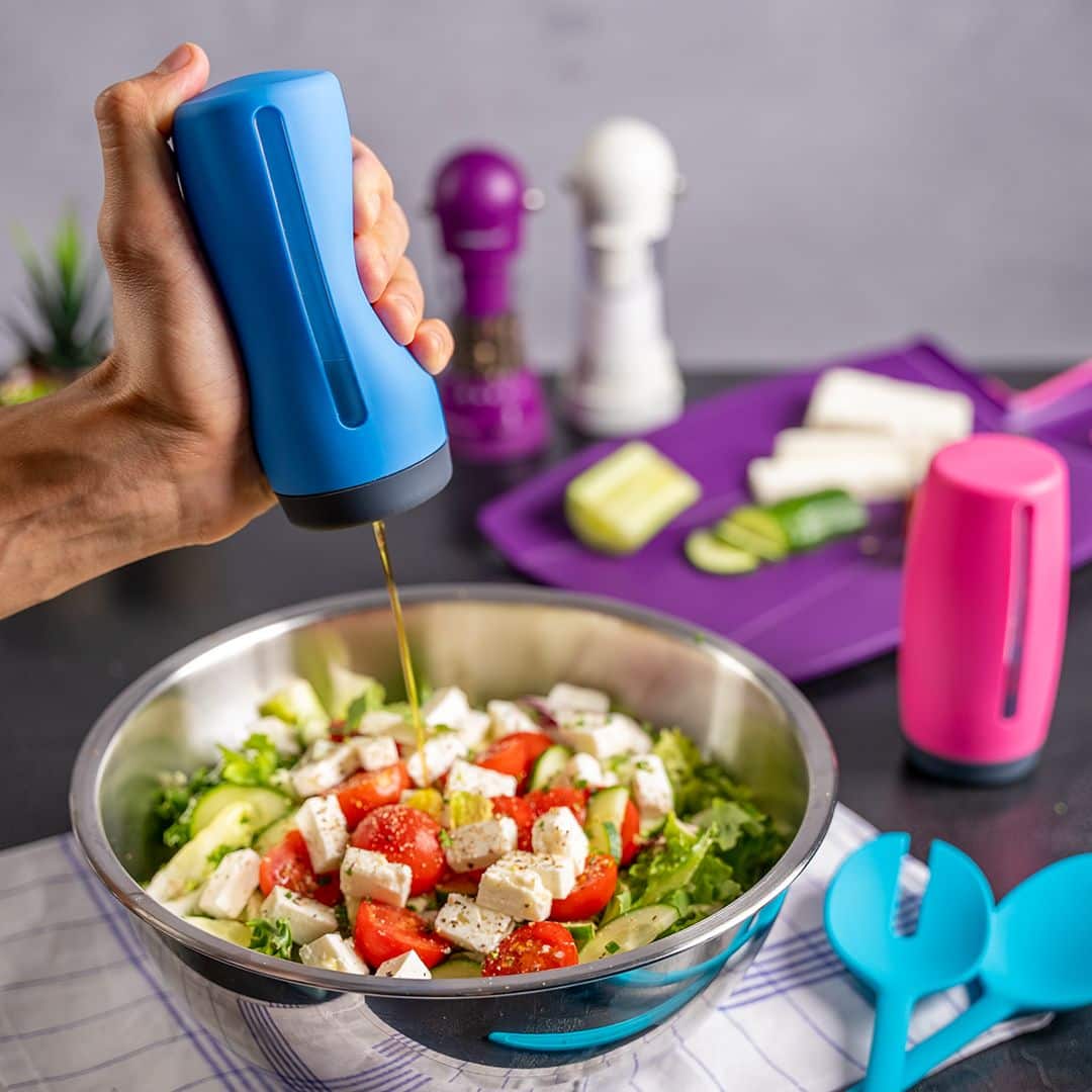 Eine Hand gibt mit dem Dosierspender der Kochblume GmbH anmutig Dressing auf einen frischen Salat in einer Schüssel, während daneben auf dem Tisch buntes Gemüse und Küchenutensilien angeordnet sind.