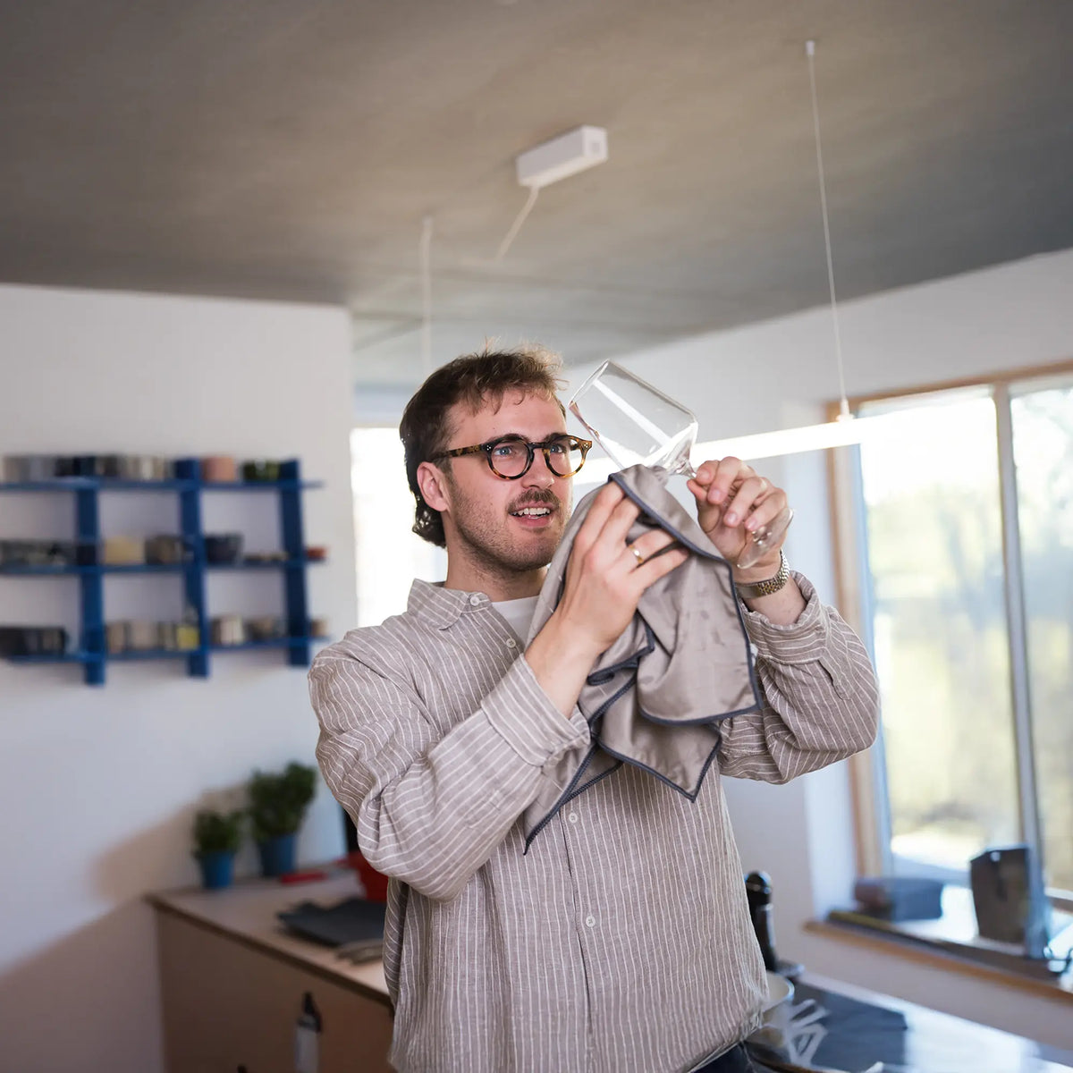 Eine Person mit Brille, die lächelt, während sie in einer modernen Küche ein Weinglas mit einem Tuch abtrocknet.