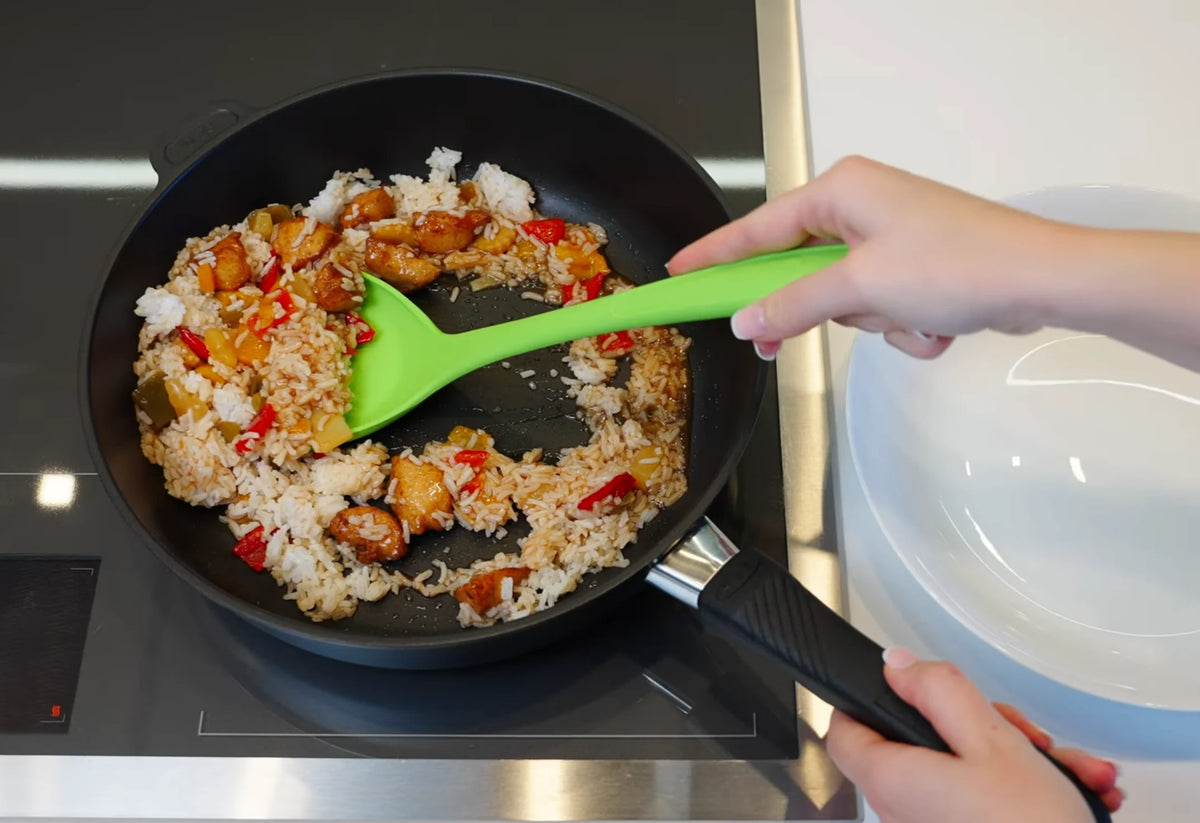 Eine Person rührt in einer schwarzen Pfanne neben einem weißen Teller mit einem grünen Pfannenwender Reis, Gemüse und Hähnchen um.