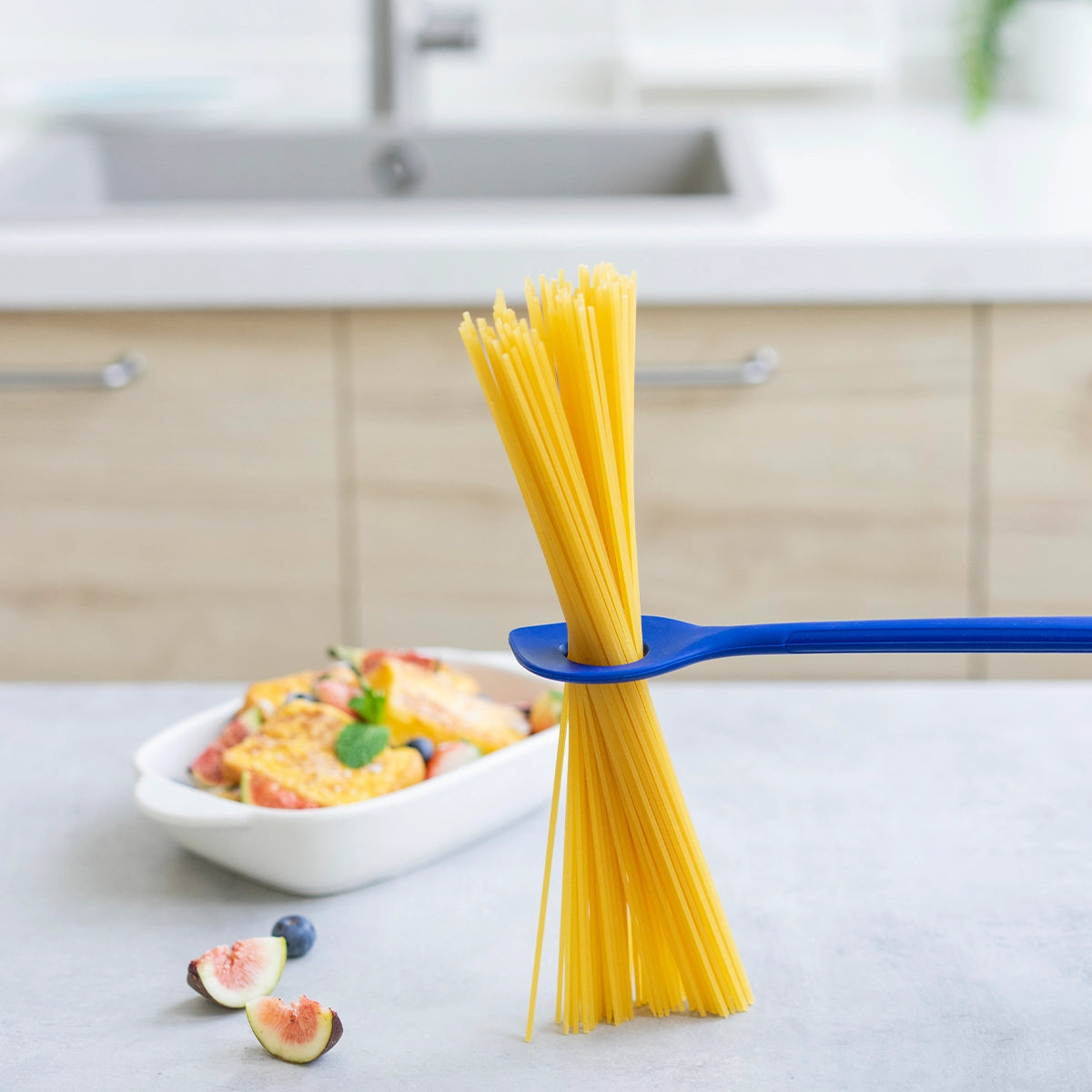 Ungekochte Spaghetti, gehalten vom Backlöffel der Kochblume GmbH, mit Feigen und einem Teller im Hintergrund auf einer Küchenarbeitsplatte – dies verdeutlicht perfekt die Essenz der stilistischen Individualisierung beim Anrichten Ihrer Zutaten, ähnlich wie die Verwendung eines Rich-Text-Editors für die präzise HTML-Formatierung Ihrer kulinarischen Präsentation.
