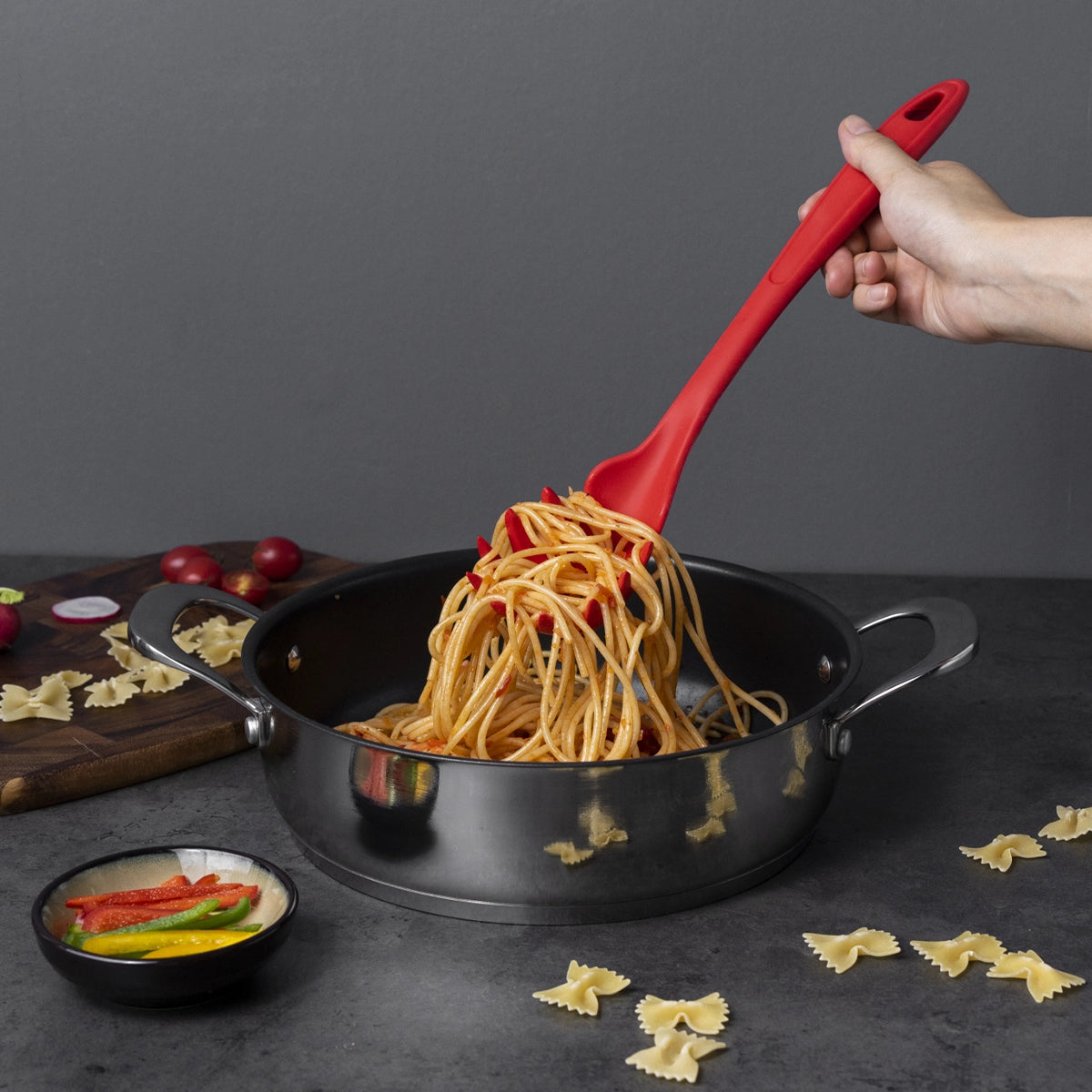 Eine Hand bedient sich des Pastalöffels der Kochblume GmbH – eines roten Spaghetti-Löffels –, um Pasta aus einem schwarzen Topf zu servieren; daneben liegen frische Pasta und Gemüse.
