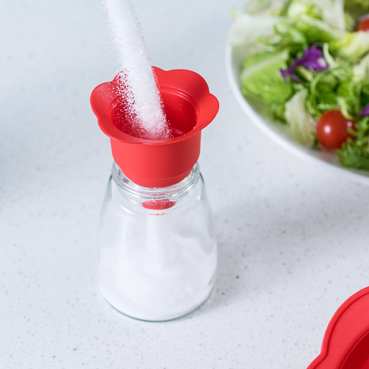 Der „Falttrichter“ der Firma Kochblume GmbH, ein roter Klapptrichter aus Silikon, wird verwendet, um Zucker in einen Glas-Shaker zu füllen; im Hintergrund ist eine Salatschüssel zu sehen.