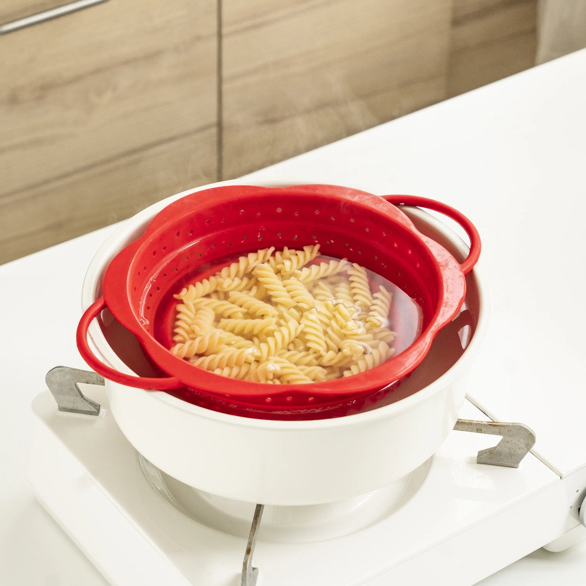 Ein rotes Faltsieb der Firma Kochblume GmbH, gefüllt mit Rotini-Nudeln, steht in einem weißen Topf mit heißem Wasser auf dem Herd und ist bereit für das perfekte Abgießen.