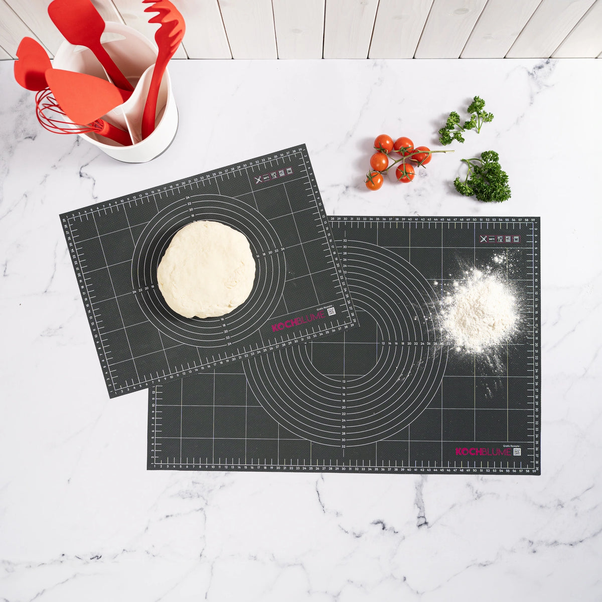Two black Gewebematte baking mats from Kochblume GmbH are shown with dough and flour, surrounded by herbs, tomatoes, and kitchen utensils. However, the product description currently only contains code instead of descriptive content. No SEO keywords could be extracted.