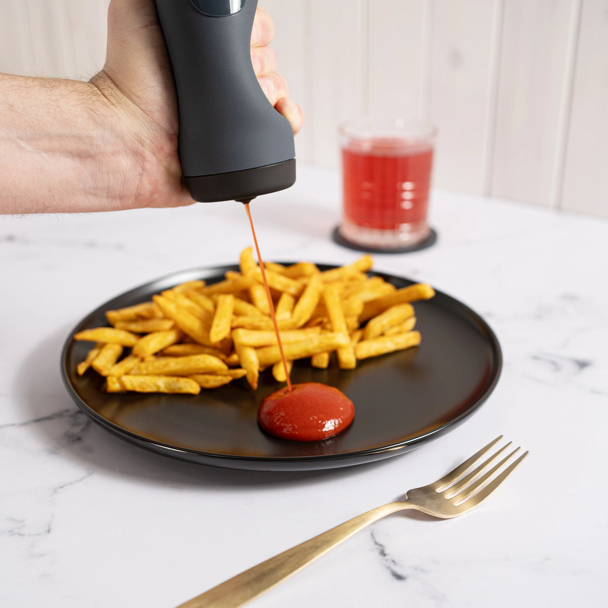 Eine Hand gibt mit dem Dosierspender der Kochblume GmbH Ketchup auf Pommes frites auf einem schwarzen Teller, daneben liegen eine Gabel und ein Glas mit einem roten Getränk.