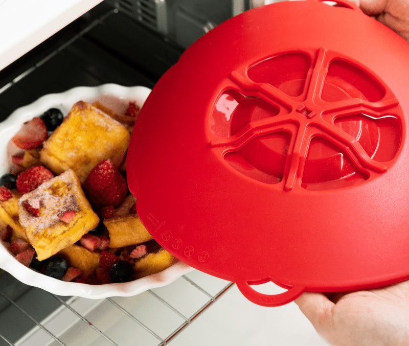 Eine Hand nimmt eine rote Silikonabdeckung von einer Auflaufform mit gebackenem French Toast und Beeren im Backofen ab.