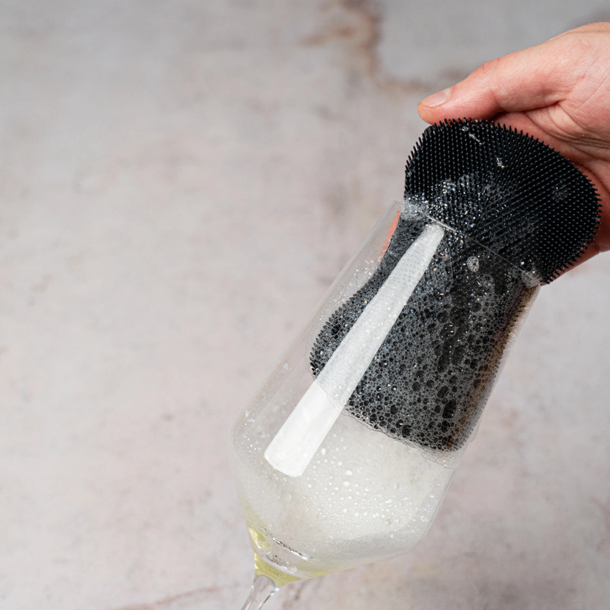 Ein Weinglas wird mit der schwarzen Silikon-Scheuerbürste „Scrubby“ der Kochblume GmbH gespült, wodurch sich im Glas Blasen bilden.