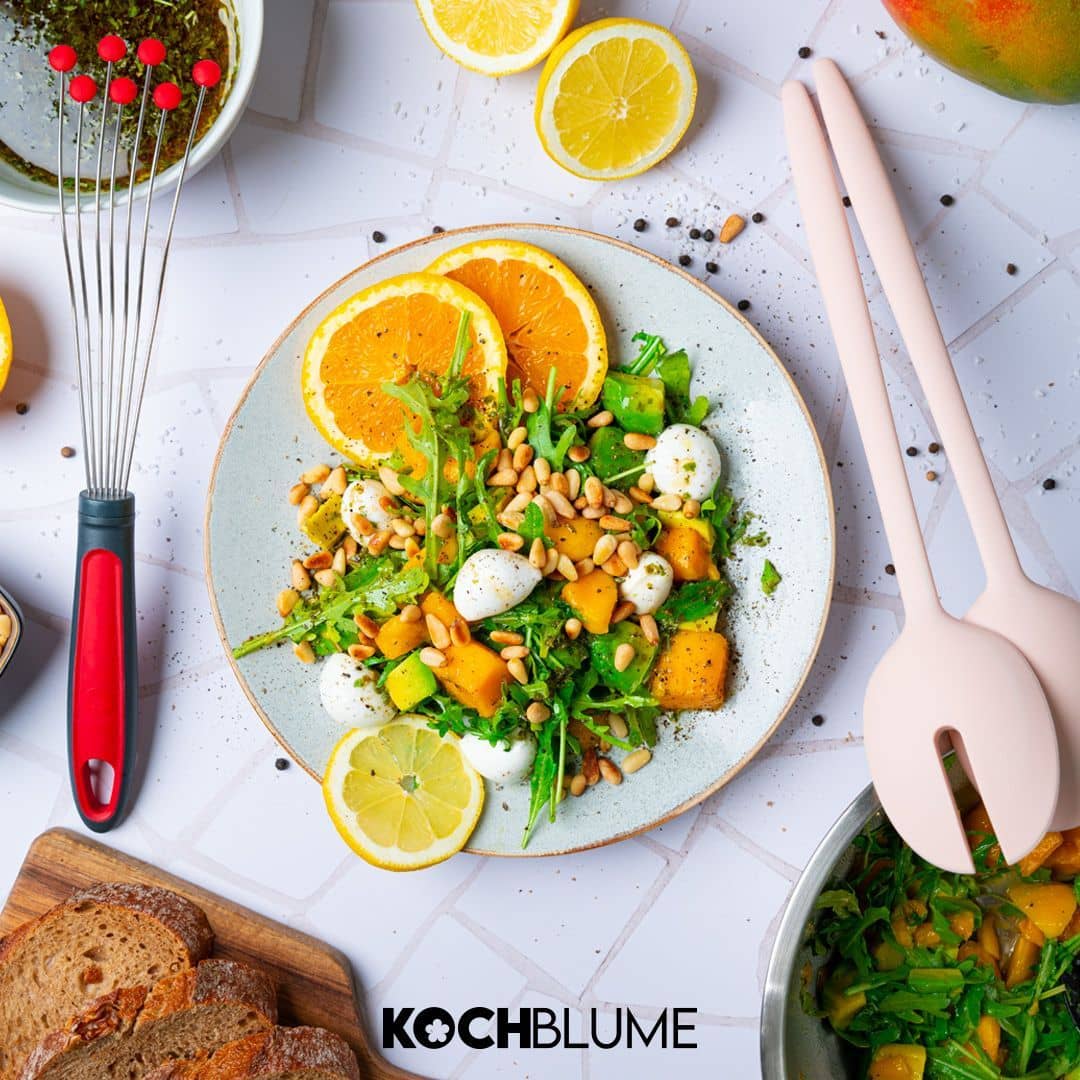 Ein frischer Salat mit Blattgemüse, Mozzarella, Mango, Pinienkernen und Orangenscheiben auf einem Teller, daneben liegt Brot.