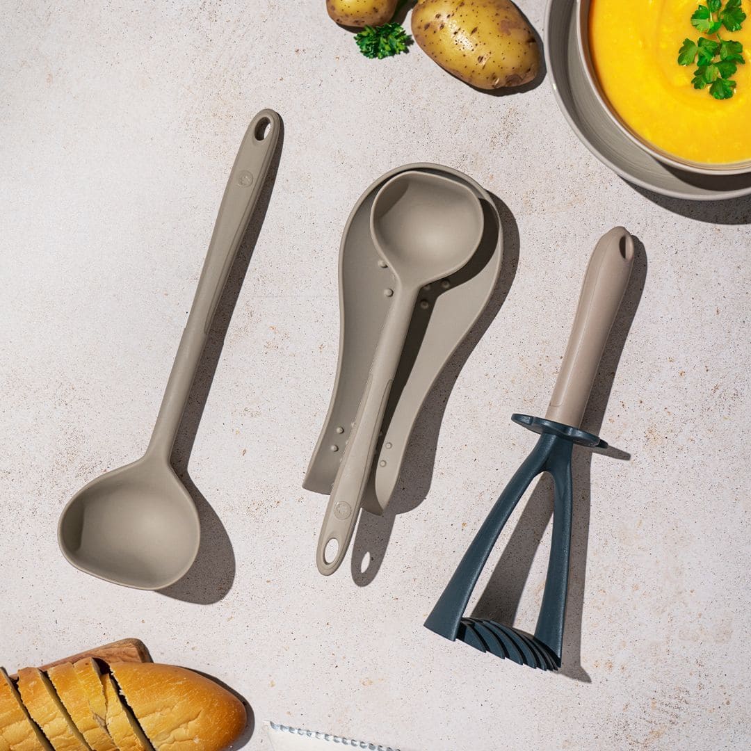 Drei beigefarbene Küchenutensilien auf einer Arbeitsplatte, daneben liegen Brot, Kartoffeln und eine Schüssel Suppe.