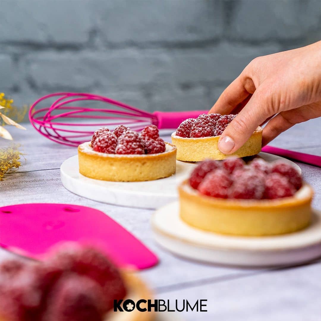 Eine Hand nimmt neben Backutensilien auf einem Tisch eine Himbeertorte in die Hand. Unten ist das Kochblume-Logo zu sehen.
