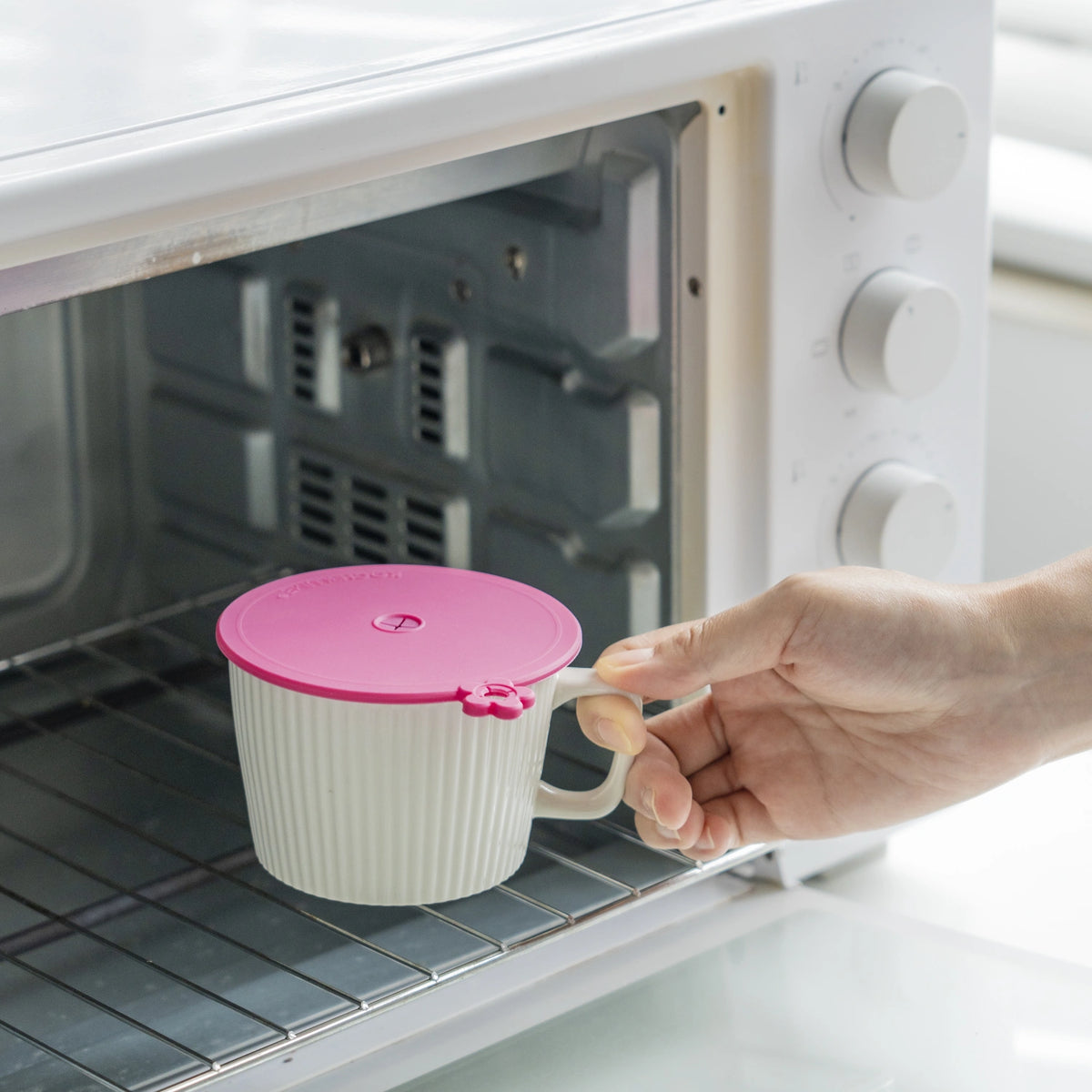 Eine Hand legt die „Glasabdeckung 3tlg.“ von Kochblume GmbH mit ihrer eleganten weißen Oberfläche und dem zartrosa Deckel in einen offenen Toaster mit Drahtgittern – ein Bild, das im klaren RTE-Stil dargestellt ist, ähnlich der Benutzeroberfläche eines Rich-Text-Editors, bei dem jede Kontur die klare Präzision eines CSS-Rahmens widerspiegelt.