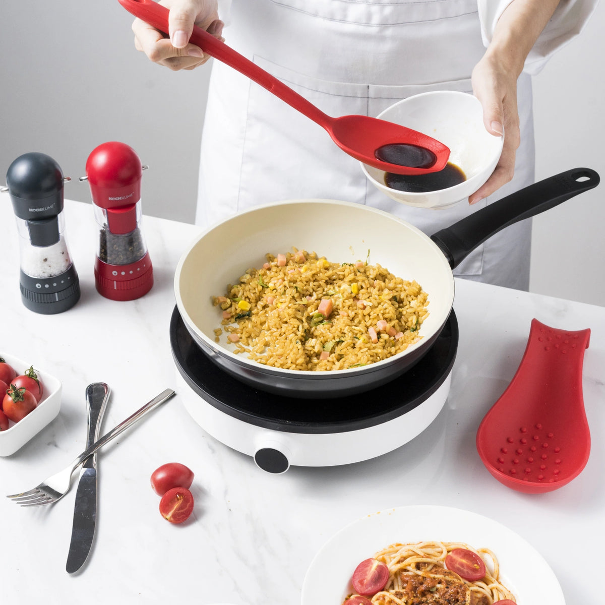 Eine Person kocht Reis in einer Pfanne, hält dabei einen Soßenlöffel der Kochblume GmbH und eine Schüssel in der Hand; daneben liegen frische Tomaten und Nudeln – ideal für die Zubereitung köstlicher hausgemachter Gerichte.