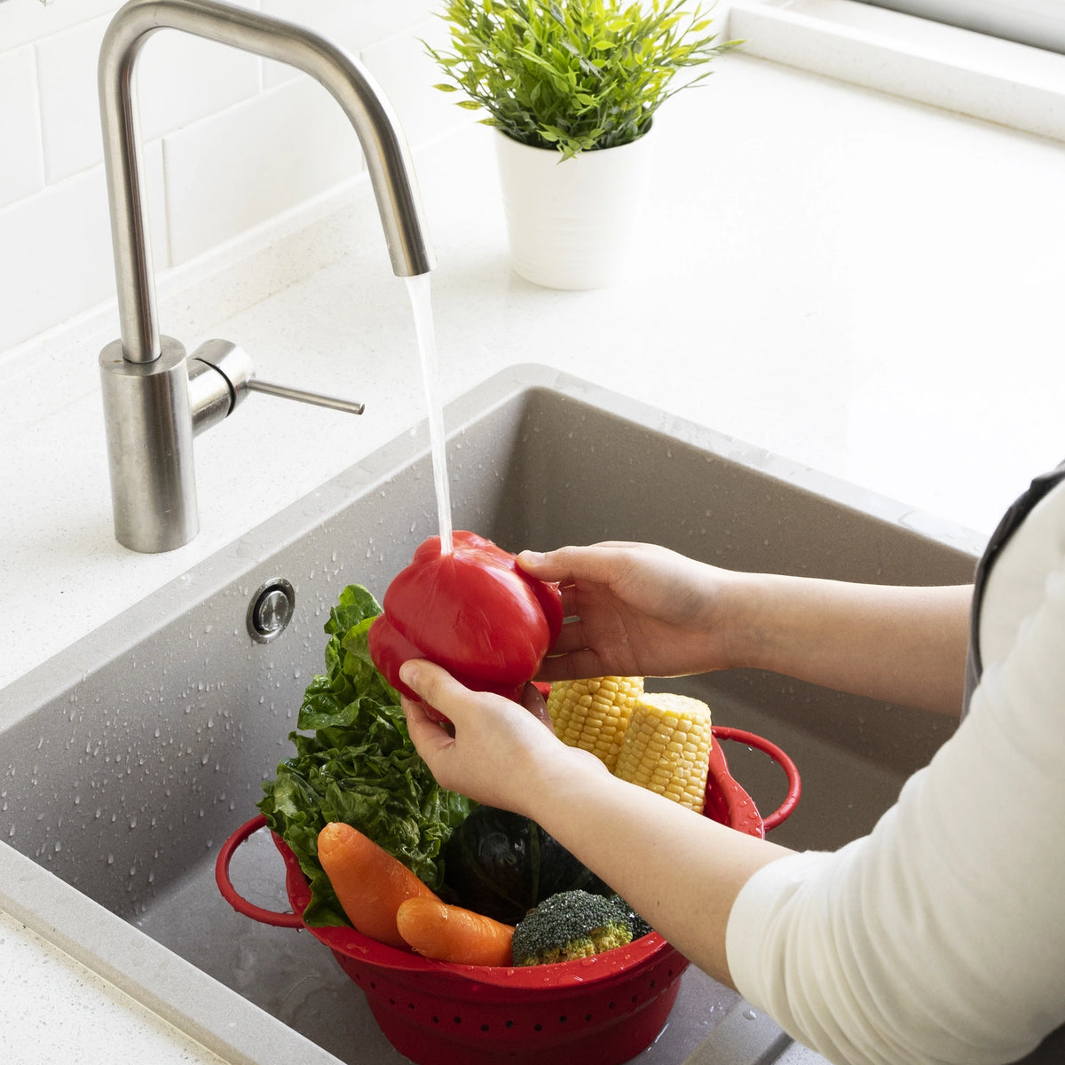 Eine Person wäscht eine rote Paprika gründlich unter dem Wasserhahn, während frisches Gemüse in einem Faltsieb der Firma Kochblume GmbH im Spülbecken liegt.