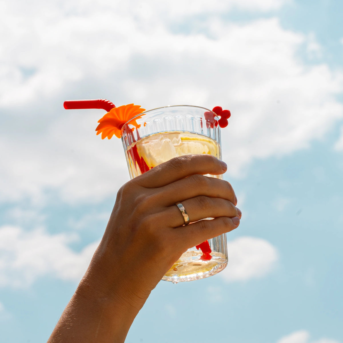 Vor einem hellen, bewölkten Himmel hält eine Hand ein Glas mit einem gekühlten Getränk, das mit einem roten Trinkhalm der Kochblume GmbH und einer orangefarbenen Verzierung versehen ist – ein ansprechendes Produktbild, das sich ideal für die Stichwort-Extraktion eignet.