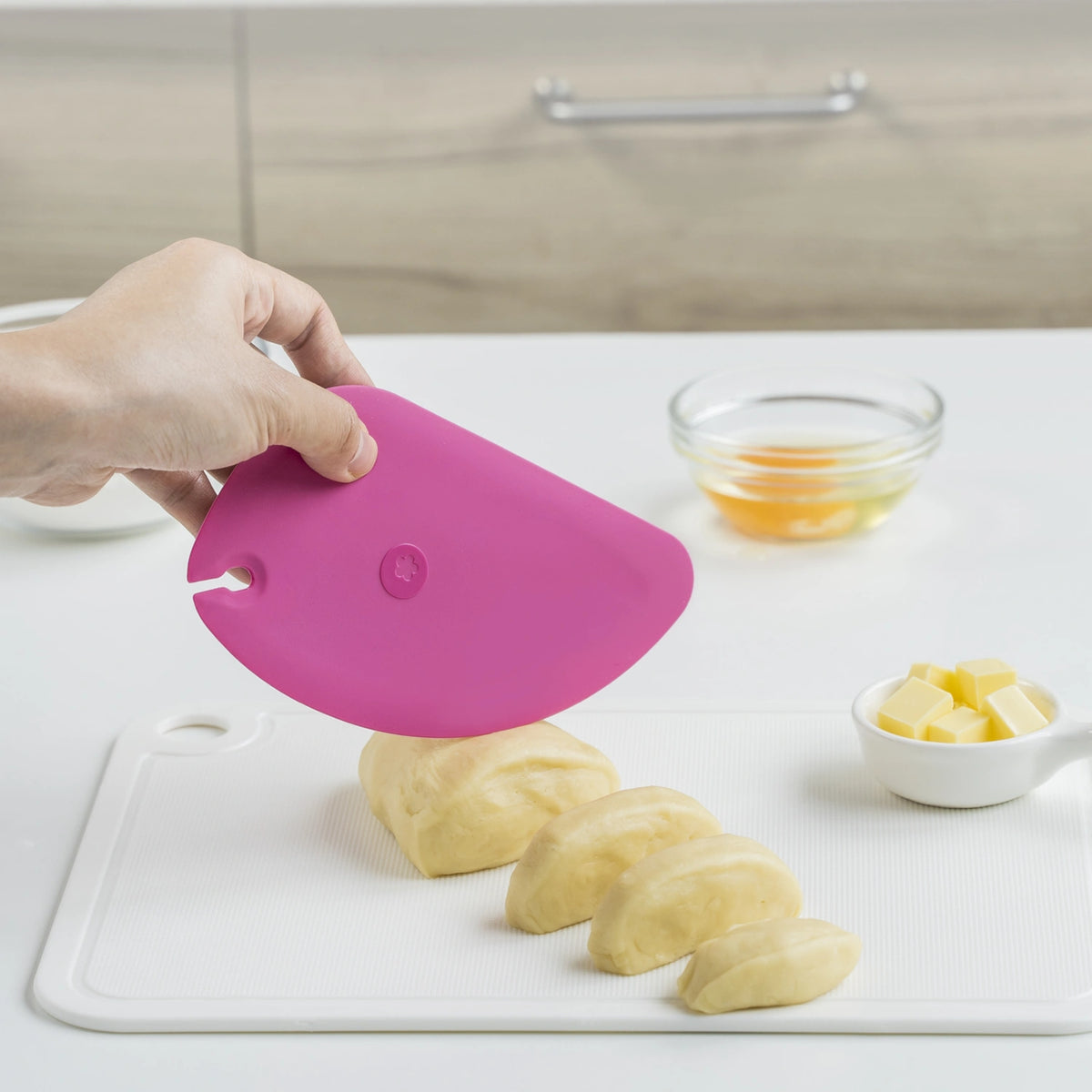 Eine Hand schneidet mit dem rosafarbenen Teigschaber „Teigkarte“ der Kochblume GmbH Teig auf einem weißen Schneidebrett, daneben liegen Butter und Eier – ein unverzichtbares Hilfsmittel für jeden Bäcker, der in der Küche auf Präzision und Komfort Wert legt.