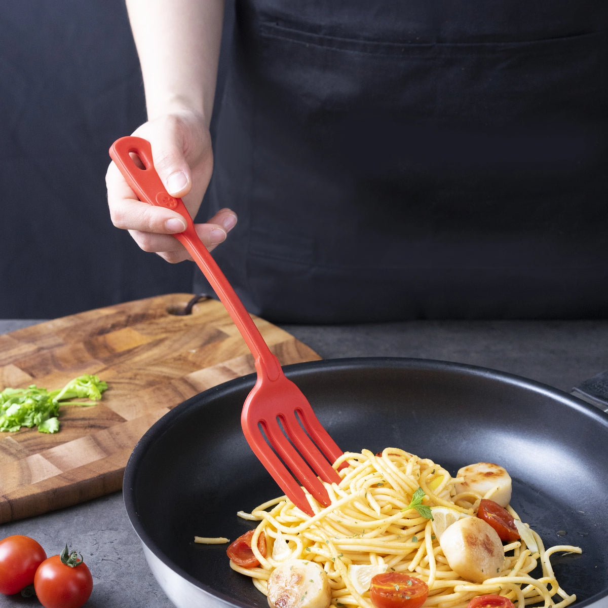 Eine Person kocht in einer Pfanne Nudeln mit Tomaten und Jakobsmuscheln und benutzt dabei die Küchengabel der Kochblume GmbH; daneben liegt ein Schneidebrett.
