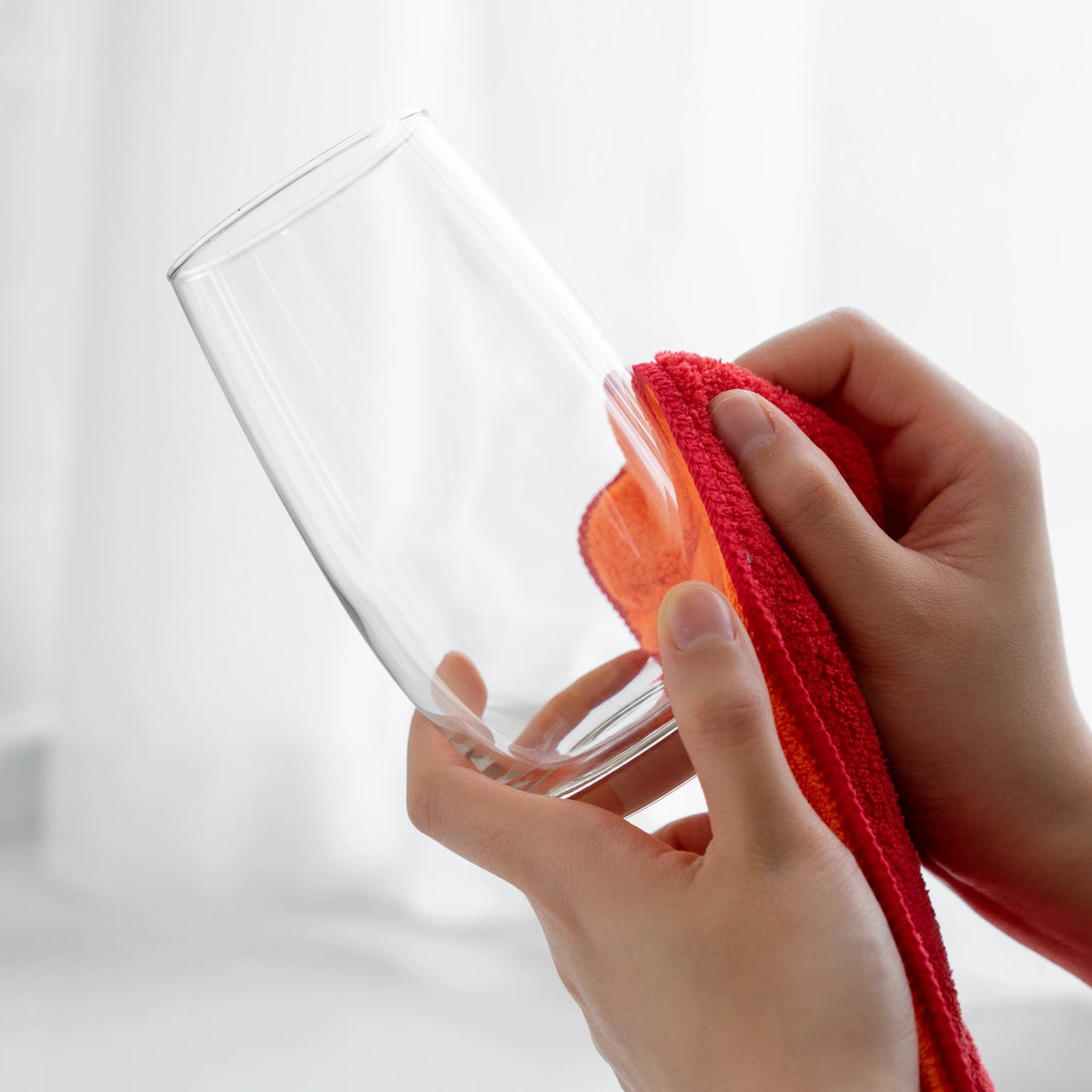 Hände, die ein klares Glas behutsam mit dem Mikrofasertuch der Kochblume GmbH vor einem weichen, hellen Hintergrund abtrocknen – eine einladende Szene, die sich perfekt dazu eignet, alltägliche Momente in Szene zu setzen.