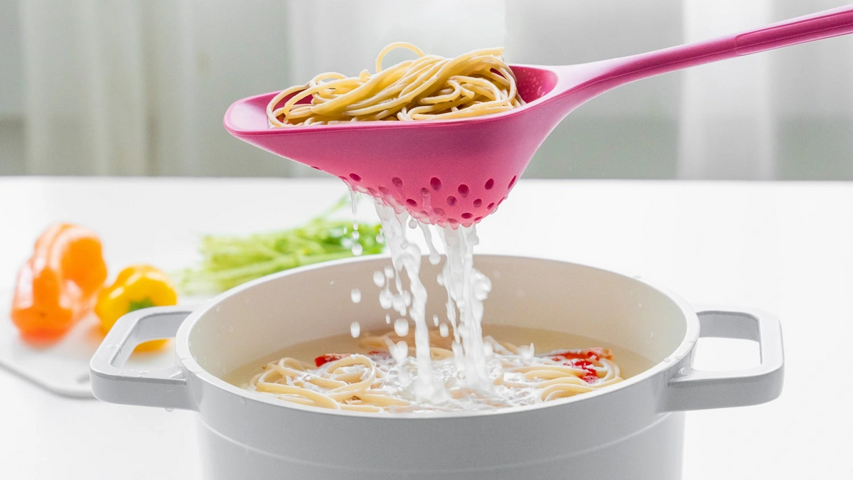 Ein rosa Schaumlöffel lässt gekochte Spaghetti über einem weißen Topf abtropfen, im Hintergrund sind verschwommene Gemüse zu sehen.