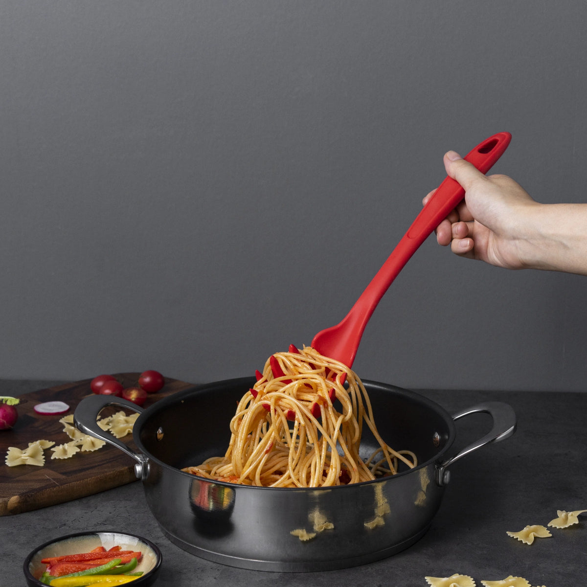Eine Hand hebt mit einem roten Pfannenwender Spaghetti aus einem schwarzen Topf auf einer dunklen Küchenarbeitsplatte.