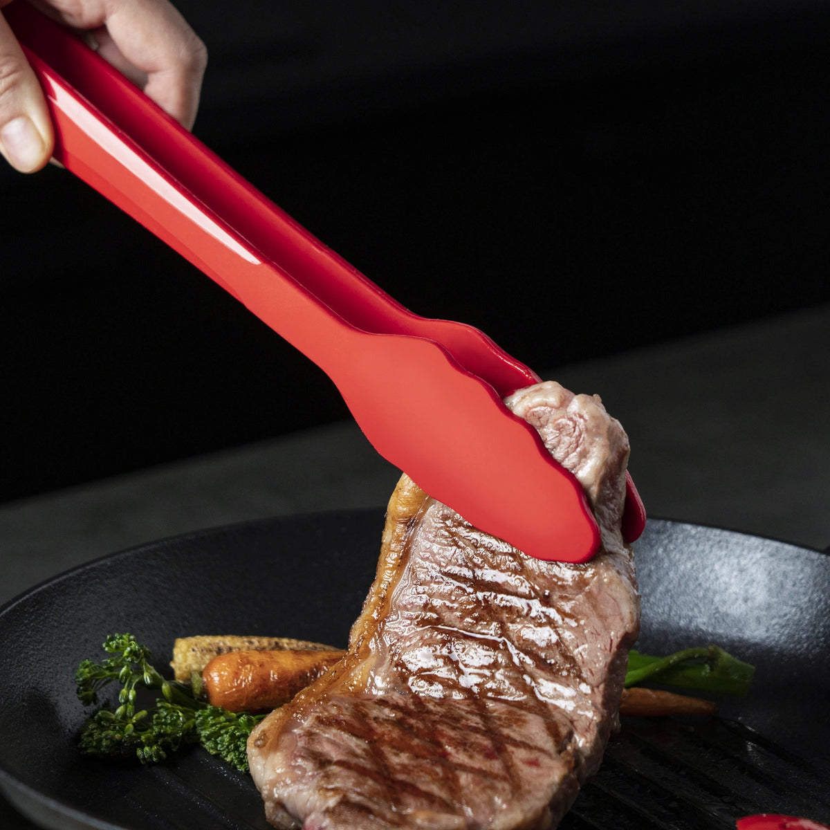 Eine rote Zange hält ein gegrilltes Steak über einer Pfanne mit Gemüse wie Möhren und Brokkoli.