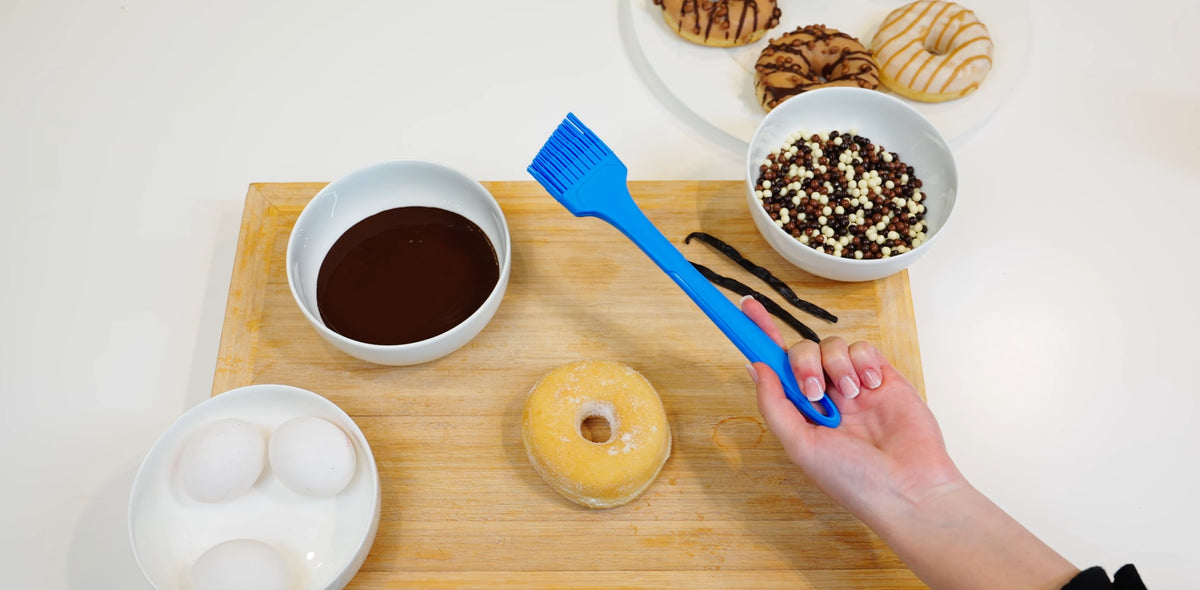 Eine Hand, die einen blauen Pinsel über einem Donut auf einem Holzbrett hält, umgeben von Eiern, Schokolade und Keksen.