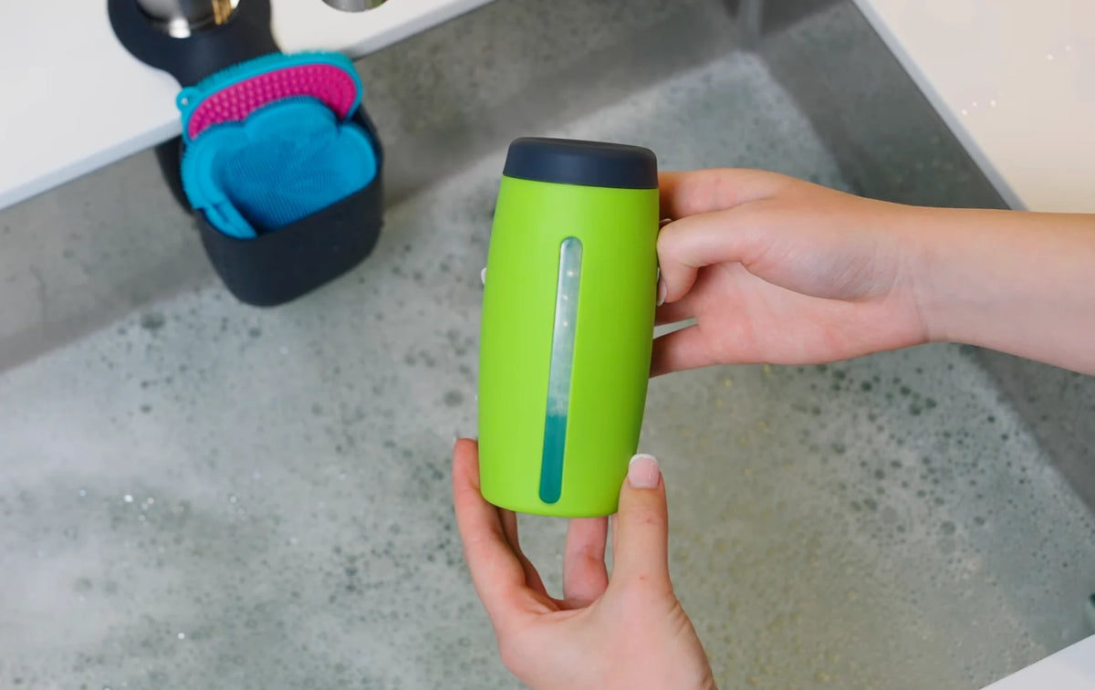 Hände, die eine grüne Flasche über ein mit Seifenlauge gefülltes Waschbecken halten, daneben steht ein schwarzer Behälter mit Reinigungsbürsten.