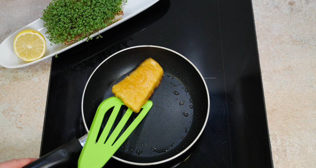 Ein grüner Pfannenwender hebt ein paniertes Fischfilet aus einer kleinen Pfanne auf dem Herd.