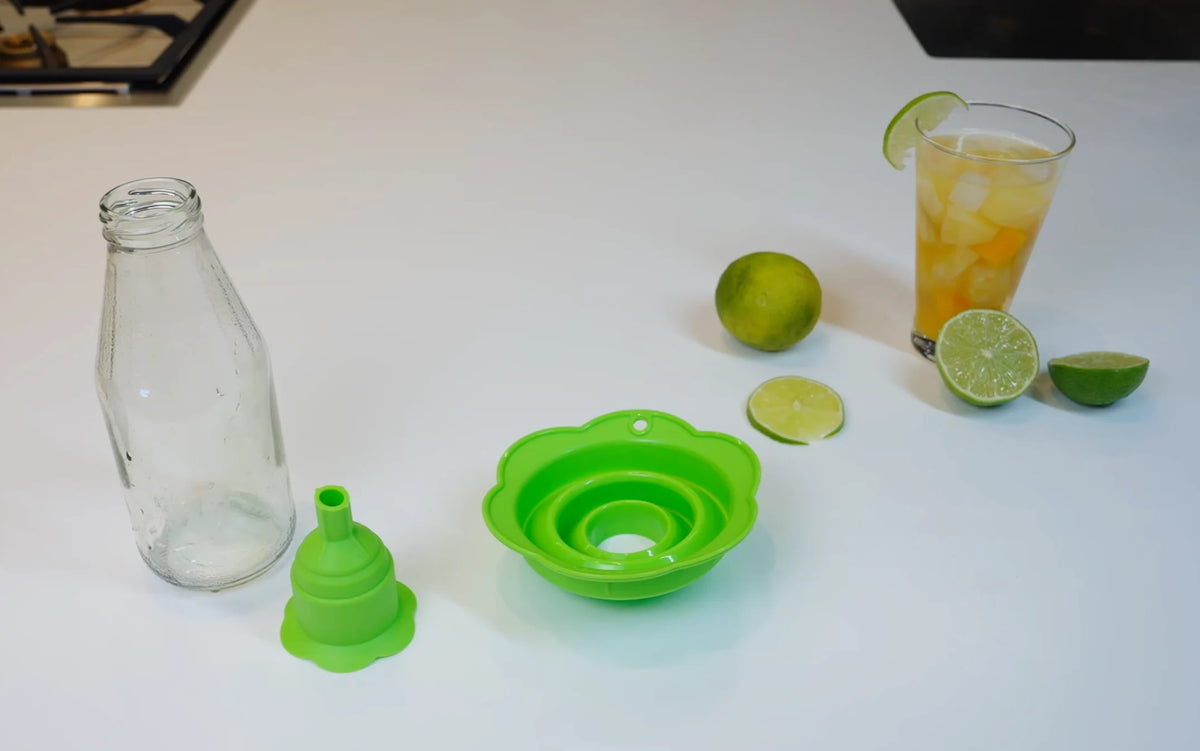 Ein grüner, zusammenklappbarer Trichter, eine leere Flasche, Limetten und ein Glas mit einem Kaltgetränk auf einer weißen Arbeitsplatte.