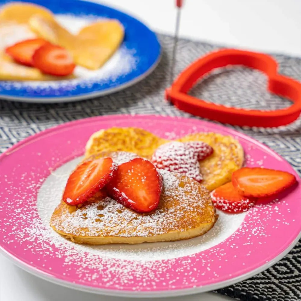 Herzförmige Pfannkuchen mit Erdbeerscheiben und Puderzucker auf einem rosa Teller.
