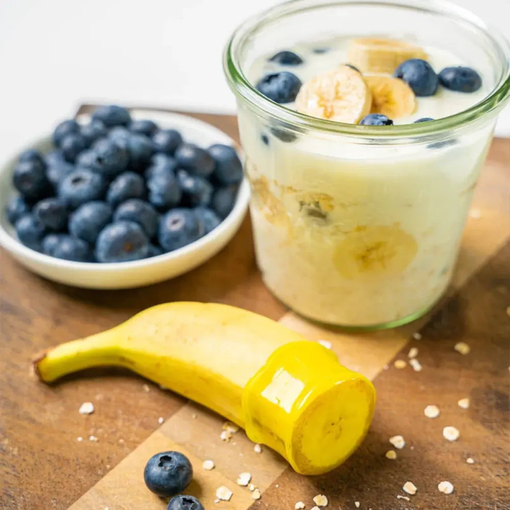 Ein Becher Joghurt mit Banane und Heidelbeeren, eine Schüssel Heidelbeeren und eine Banane auf einem Holzbrett.