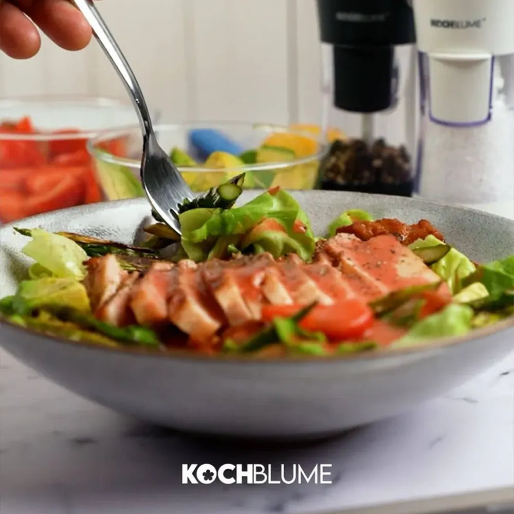 Ein Gabelstapler hebt einen Salat mit angebratenen Fleischscheiben aus einer Schüssel; im Hintergrund sind frisches Gemüse und Gewürze zu sehen.