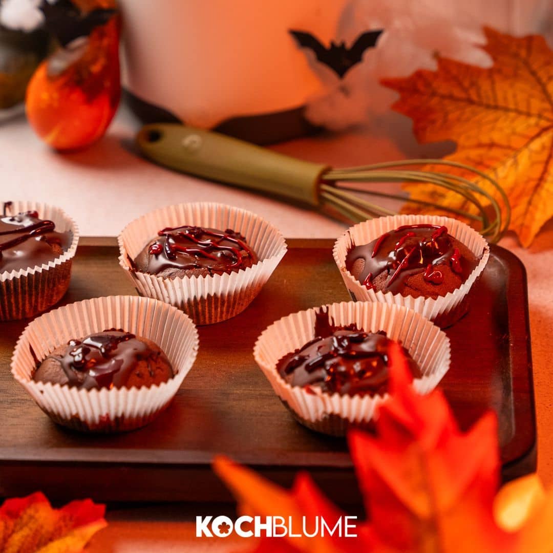 Fünf Schokoladen-Cupcakes in Papierförmchen auf einem Tablett, umgeben von Herbstblättern und Halloween-Dekorationen.