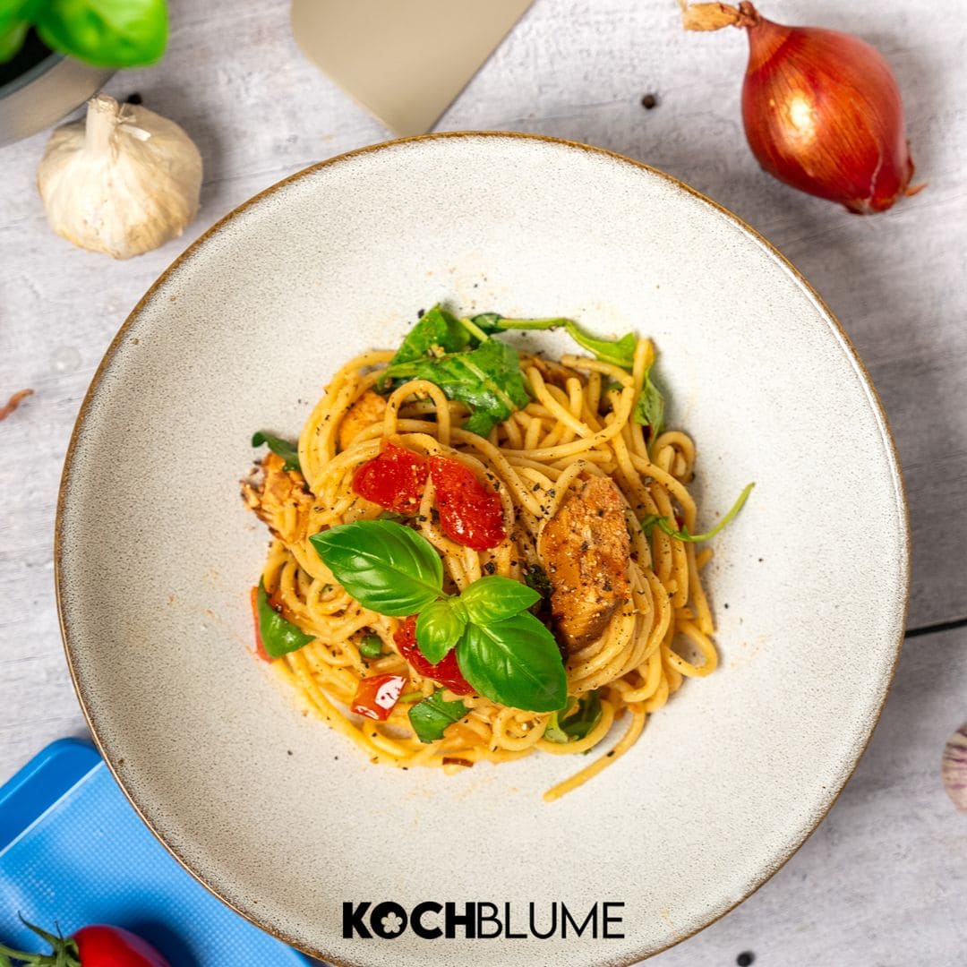 Nudeln mit Tomaten, Hähnchen, Spinat und Basilikum, serviert in einer weißen Schüssel auf einem hellen Tisch.