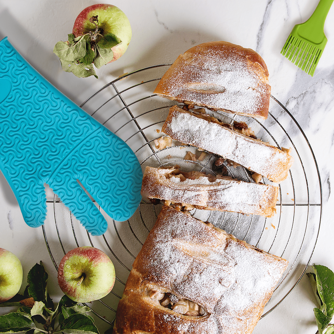 In Scheiben geschnittener Apfelstrudel auf einem Kuchengitter, daneben liegen Äpfel, ein blauer Ofenhandschuh und ein grüner Backpinsel.