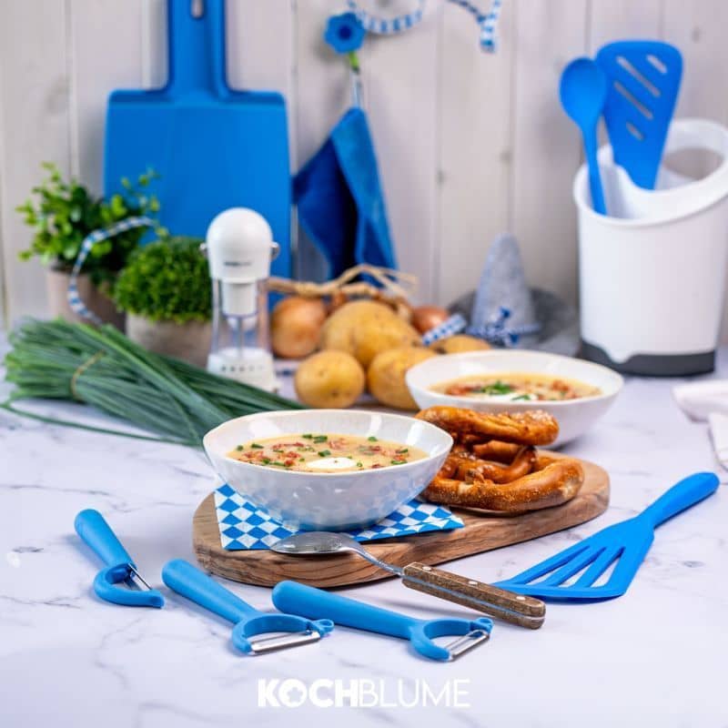 Zwei Suppenteller mit Brezeln, Kartoffeln, Schnittlauch und blauen Küchenutensilien auf einem weißen Tisch.