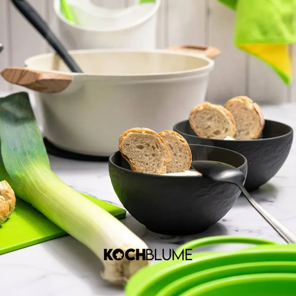 Zwei schwarze Felix-Schüsseln, gefüllt mit Suppe und Brot, ein Lauch und Kochgeschirr auf einer Küchenarbeitsplatte. Im Vordergrund das Logo der Kochblume GmbH.